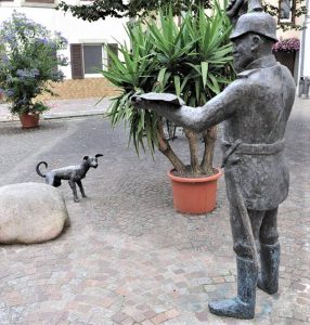 DER AMTSBOTE in Hornberg, Bronze. Auftrag der Sparkasse Haslach-Hornberg. Der Polizist (Brüstle Bollis) mit dem Strassenköter sollte an einer beliebten Persönlichkeit der Stadt Hornberg erinnern, welcher in der Silvester Nacht oben auf dem Schloß zur großer Freude der Bürger Trompete spielte, um das Neues Jahr anzukündigen. Nun, eine Pickelhaube hat er nie getragen; aber Auftraggeber haben manchmal seltsame Einfälle...